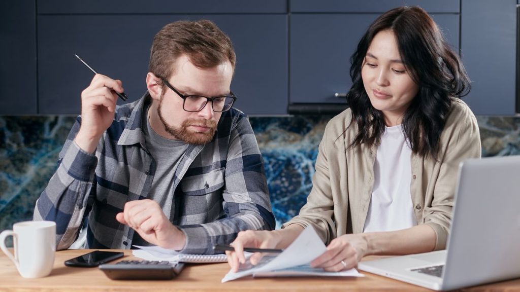 زوجان يراجعان أوراق الميزانية وحسابات المنزل بجانب آلة حاسبة وحاسوب محمول